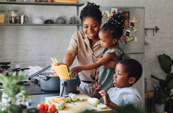 A woman holding her daughter and cooking with her children in the kitchen.